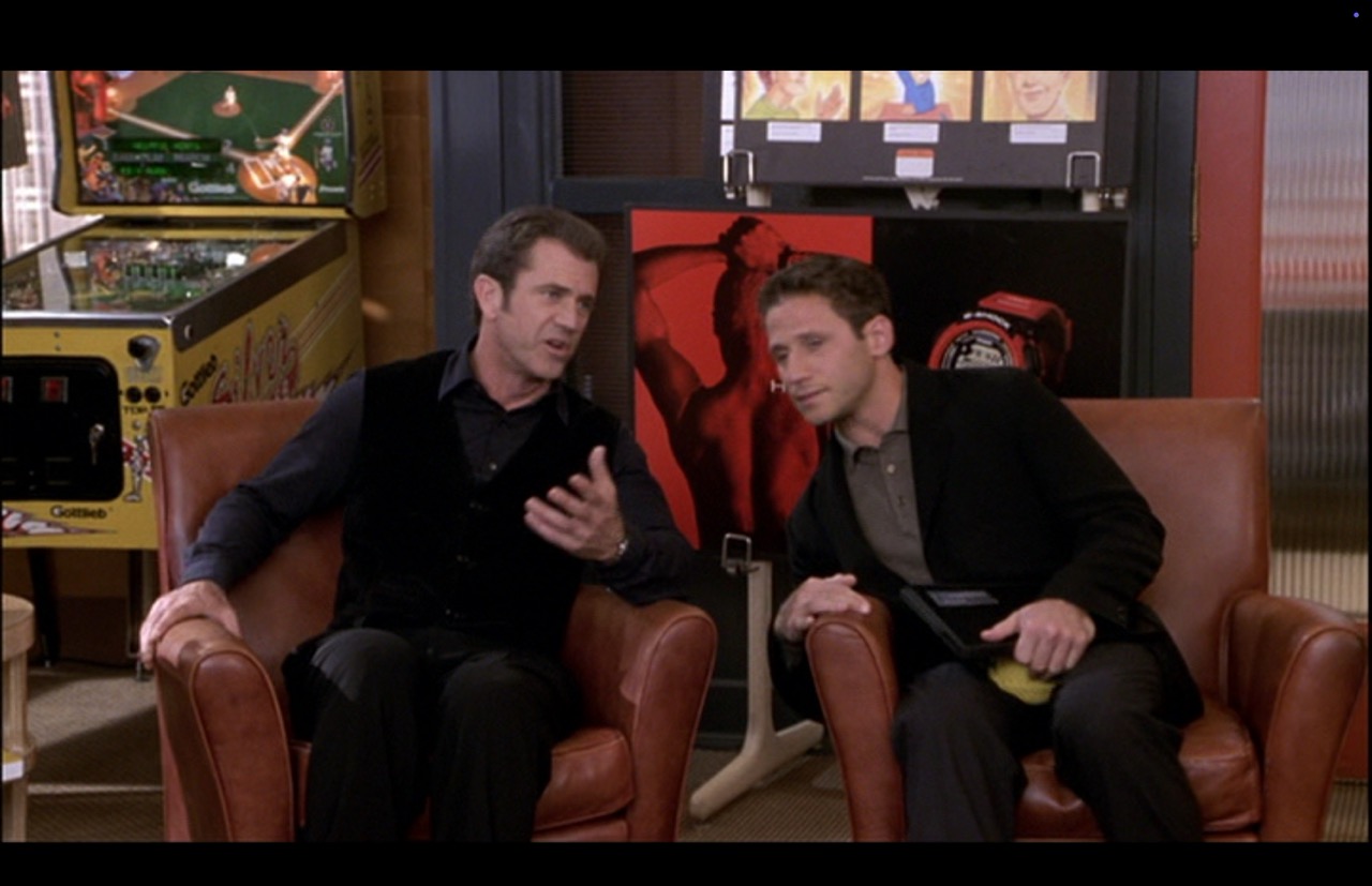 Nick (Mel Gibson) takes his seat in front of the Silver Slugger, sitting next to Morgan (Mark Feuerstein), his friend/co-worker/confidant. The Silver Slugger is powered up and its lights are on.