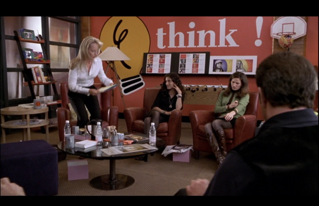 Darcy (Helen Hunt) is about to take her seat on the opposite side of the room, which is decorated with large colorful creativity-inspiring graphics and a foosball table, in front of which sit two executives. A basketball hoop hangs on the wall behind and above them, off to the side.