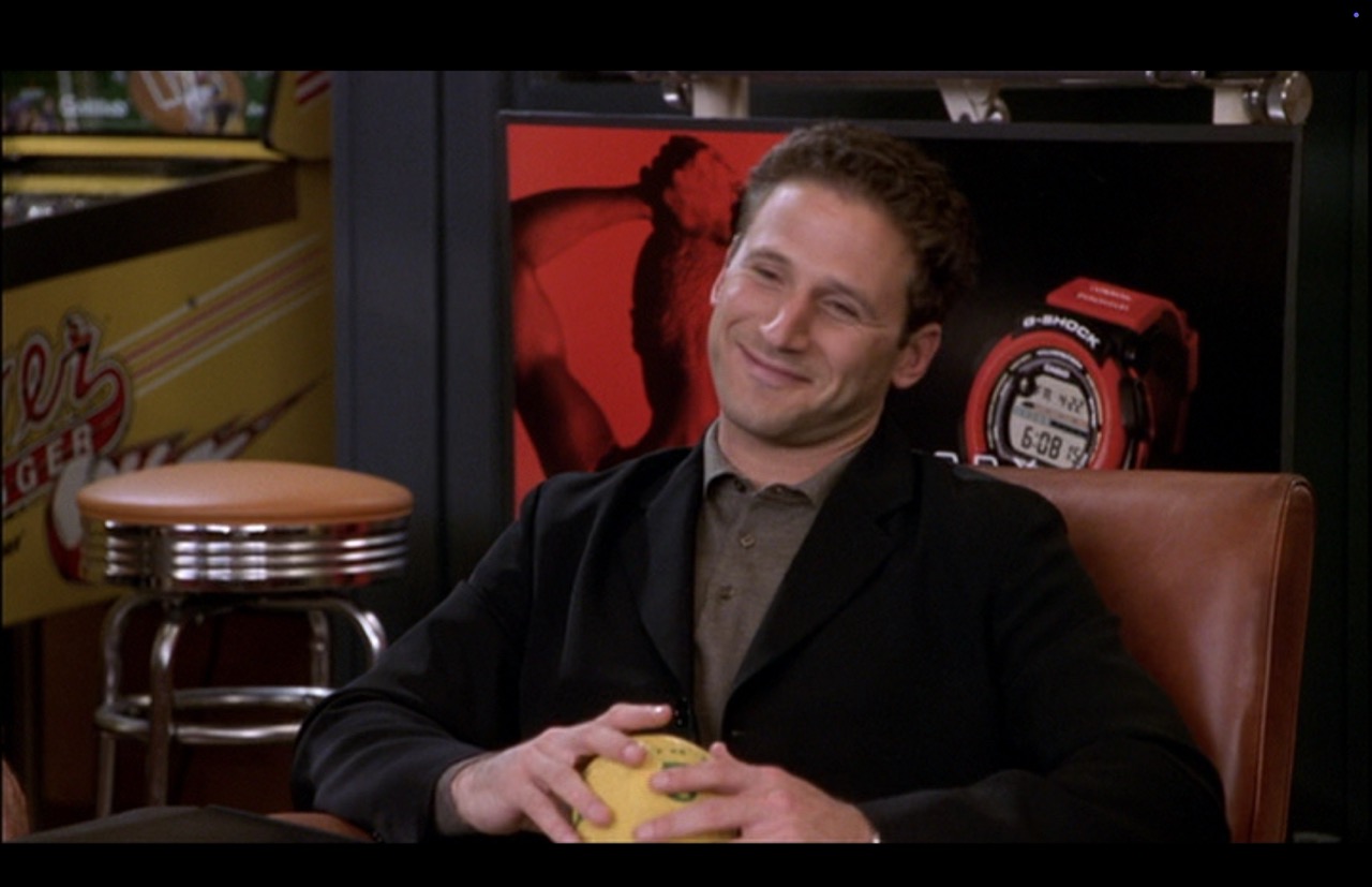 Morgan reacts to dialogue. He is alone in the frame in a medium shot with the Silver Slugger and a chrome diner stool dominating the left-most quarter of the image.
