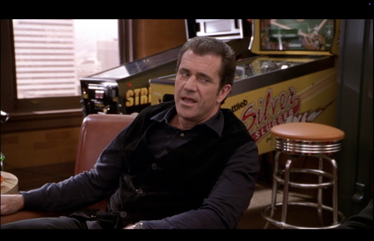 The camera’s frame centered back on Nick in a medium shot. The Silver Slugger fills the space between the co-workers. The side graphics “Silver Slugger” and nearly all of the word “Gottlieb” are in soft focus, noticable and legible.