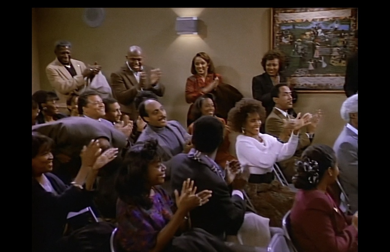 The scene begins. The audience in the church’s large activity room applauds happily in reaction to some just-completed performance. Standing against the wall on the side of the room, Roc (Charles S. Dutton) and wife Eleanor (Ella Joyce) are also enthusiastically clapping. On that wall, closer to the stage, is the framed poster “Church Picnic,” lit from above by a wall-mounted picture light. The framed poster takes up about one twelfth of the image.