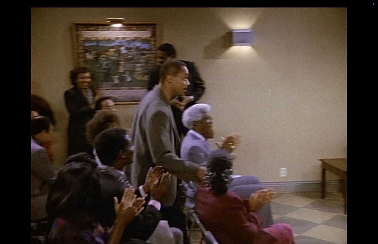 Roc’s brother Joey (Rocky Carroll) strides down the center aisle to the stage to direct his trumpet students in their perfomance. The “Church Picnic” poster takes up about one-twelfth of the screen. The poster is motion blurred. The only other items on the gray-beige wall is a sconce throwing light upward and downward, a motion-blurred electrical wall outlet, and a baseboard. Much of the wall is a large, undecorated expanse.