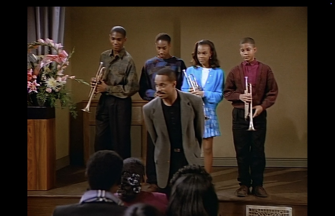 Joey introduces his four trumpet students on stage at the front of the hall. The wall behind them is essentially blank. A large pastel bouquet of flowers decorates the viewer’s left.
