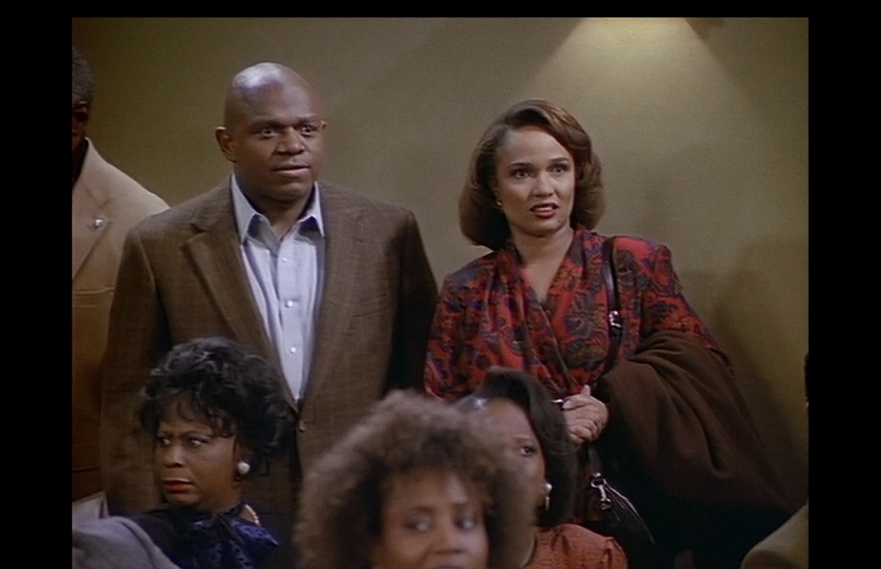A medium shot framing Roc and Eleanor in front of blank wall shows the beginnings of dismay on their faces as the trumpets wail and squeak cacophonously.