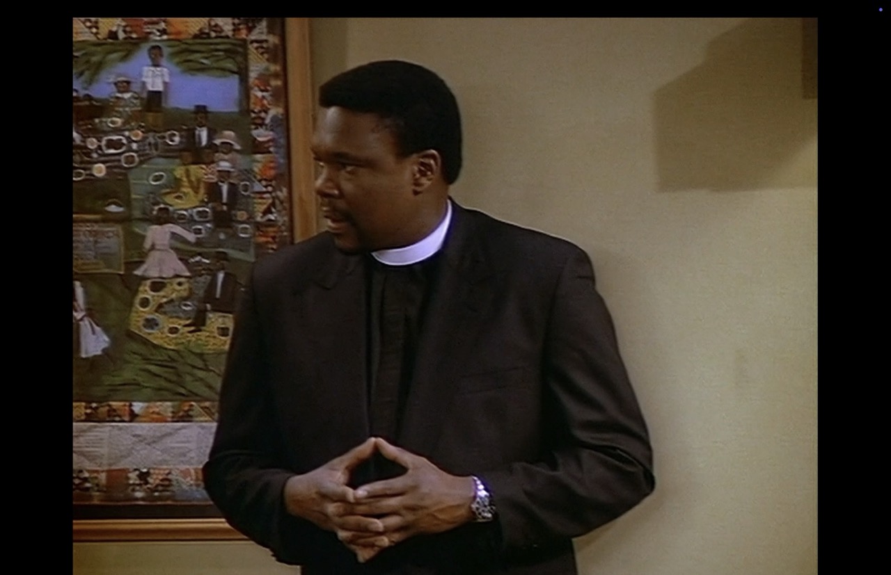 Reverend Hill (Harold Sylvester) is standing beside the same wall as Roc and Eleanor, though closer to the stage. Hill is framed by himself as he looks back toward Roc and Eleanor showing concern. The “Church Picnic” poster fills about one-third of the screen and is in focus. The artwork’s figures and their plates of food can be made out.