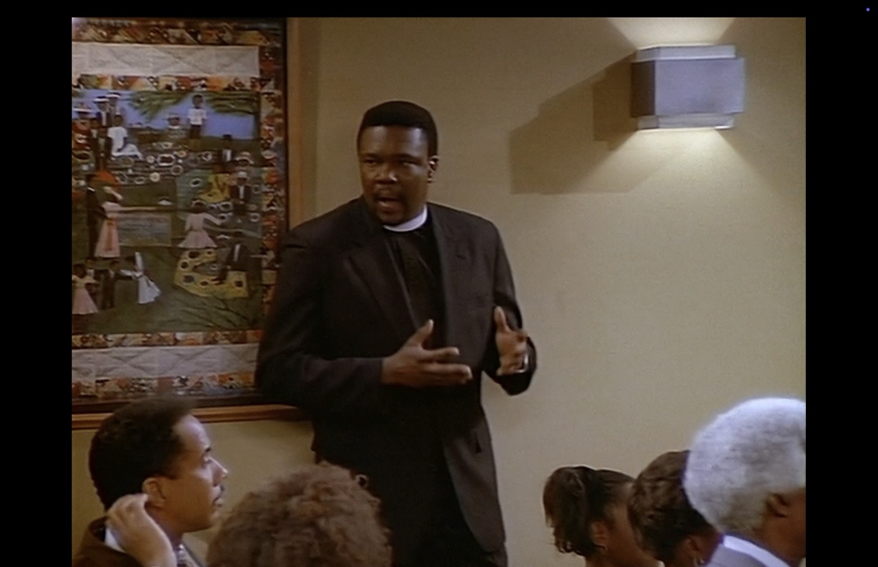 While looking back at Roc, Hill begins to applaud the now completed performance. The “Church Picnic” poster dominates the left third of the image; considerable detail can be seen.