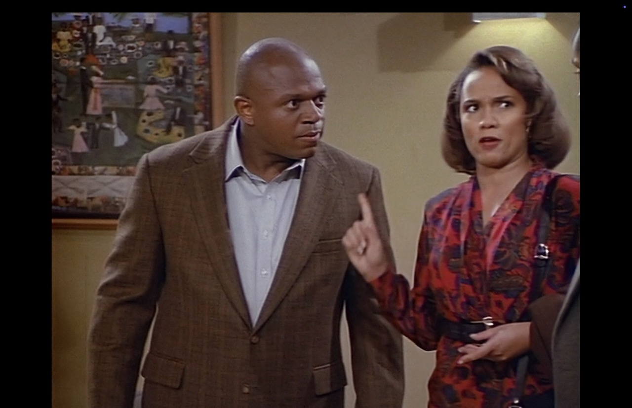 Now moved toward the hall’s front to confront Joey, Roc leans and stares at his brother while Eleanor angrily waves a finger in a medium shot that leaves Joey mostly out of frame. A portion of the poster, appearing behind Roc’s right shoulder, fills approximately one-sixth of the background.