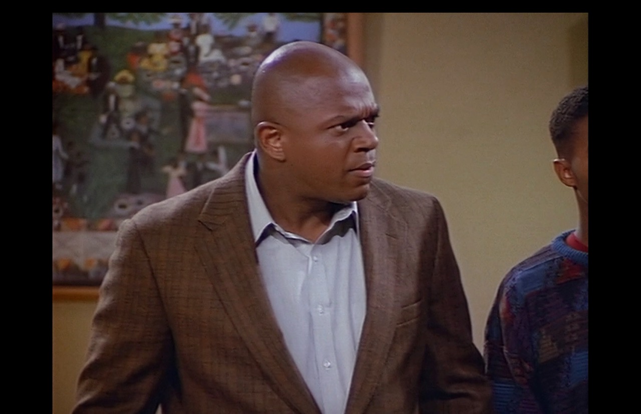 A medium close-up on Roc shows his growing disbelief as Joey is offered improbable compliments on his music teaching from implacably proud parents. The poster, out of focus, dominates the left half of the background.