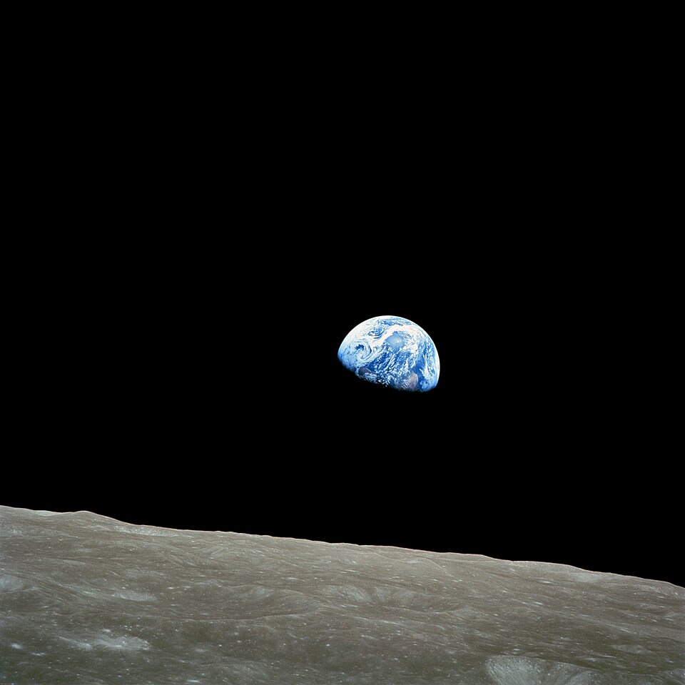 In the featureless black of space, the earth appears rising over the lunar surface which spreads out in the foreground
